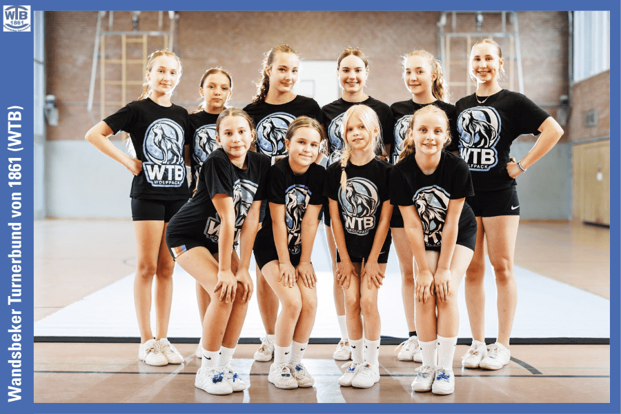 Gruppe von zehn Mädchen in T-Shirts mit WTB-Logo, lächelnd in einer Turnhalle auf einem Sportboden posierend.