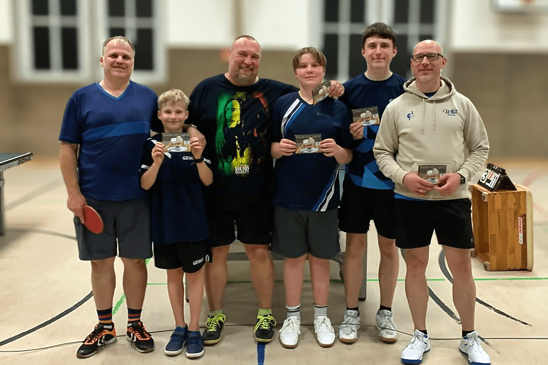 Fünf Personen in Sportkleidung halten Pokale, stehen auf einem Tischtennisplatz und lächeln in die Kamera.