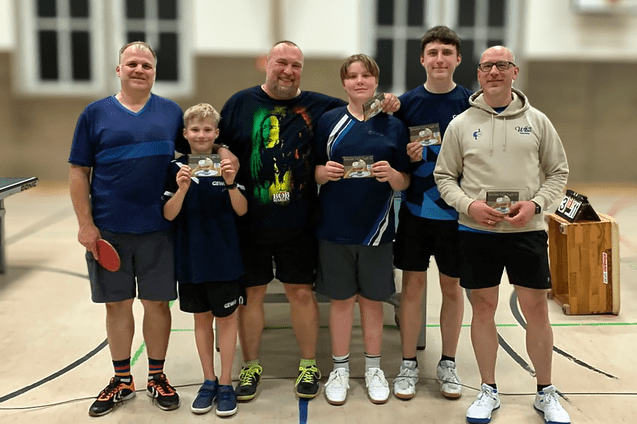 F&uuml;nf Personen in Sportkleidung halten Pokale, stehen auf einem Tischtennisplatz und l&auml;cheln in die Kamera.