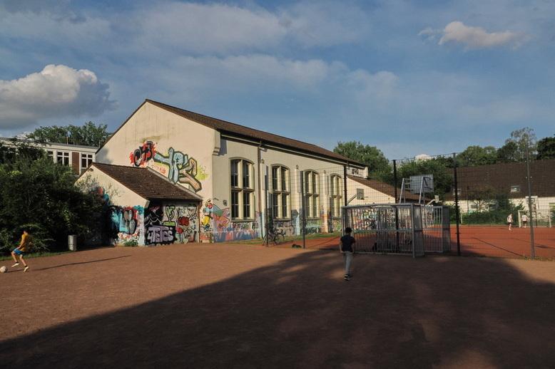 Graffiti-bedeckte Schule neben einem Sportplatz, auf dem Kinder spielen, unter einem blauen Himmel mit Wolken.