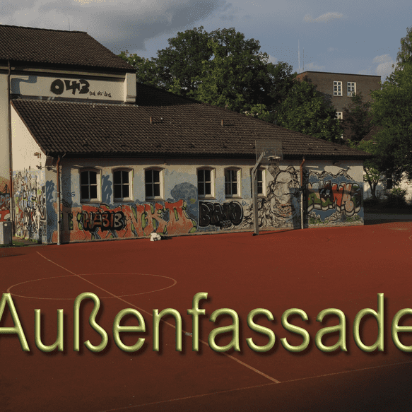 Graffiti-bedeckte Au&szlig;enfassade eines Schulgeb&auml;udes an einem roten Sportplatz, umgeben von B&auml;umen.