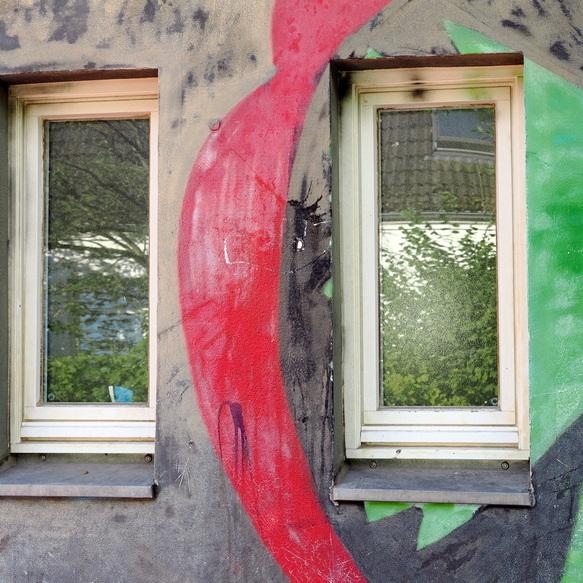 Zwei Fenster mit reflektierenden Scheiben, umgeben von bunten Graffiti auf einer Wand.