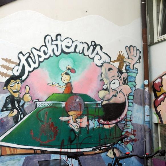 Wandmalerei mit Tischtennis-Motiv: Spielerin und Zuschauer, Schriftzug &bdquo;Tischtennis&ldquo; &uuml;ber einem Tischtennistisch.