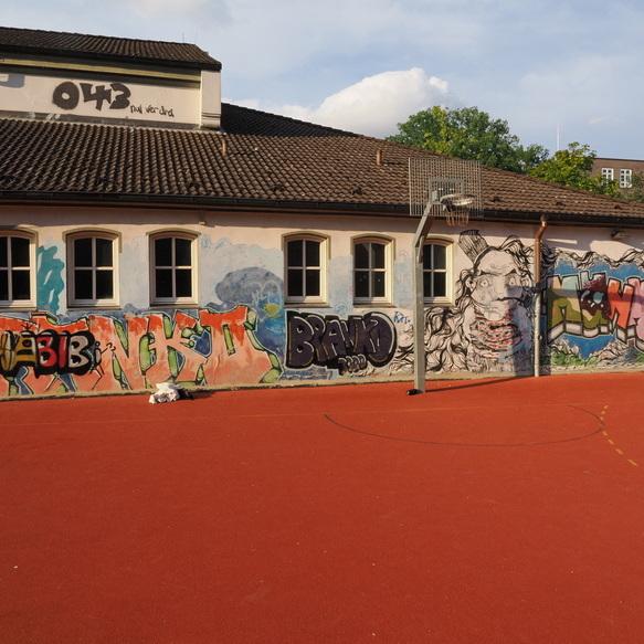 Graffiti-Wand an einem Schulgeb&auml;ude mit verschiedenen Schriftz&uuml;gen und Portr&auml;t, im Vordergrund ein Basketballkorb.