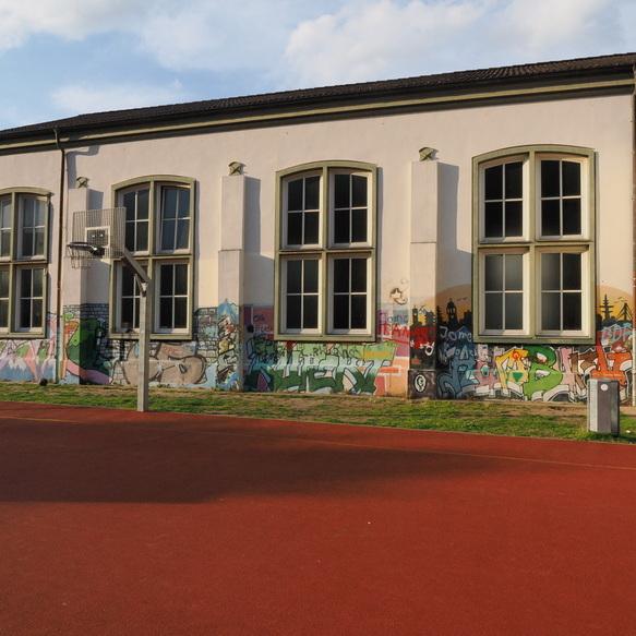 Graffiti verzierte Wand eines Schulgeb&auml;udes mit zwei gro&szlig;en Fenstern, davor ein Basketballkorb auf einem roten Platz.
