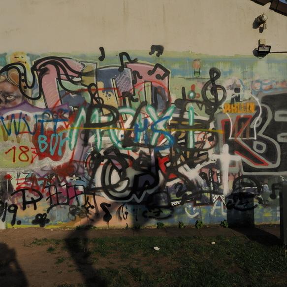 Wand mit buntem Graffiti, das Musiknoten, Schriftz&uuml;ge und abstrakte Formen zeigt, im Freien neben einem Sportplatz.