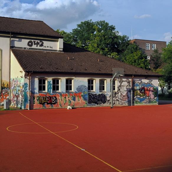 Graffiti-bedeckte Wand eines Schulgeb&auml;udes neben einem Basketballfeld mit rotem Belag, umgeben von B&auml;umen.