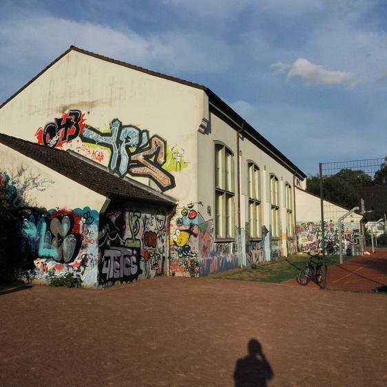 Graffiti-bedeckte Wand eines Geb&auml;udes neben einem Tennisplatz, im Hintergrund eine Tennisnetz und B&auml;ume.