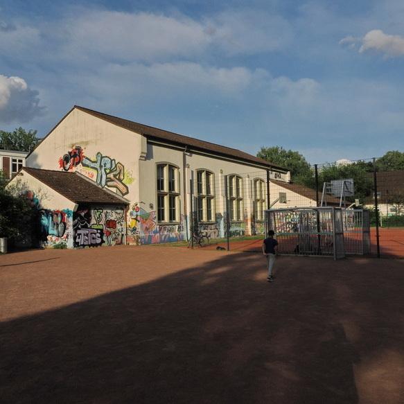 Graffiti-bedeckte Schule neben einem Sportplatz, auf dem Kinder spielen, unter einem blauen Himmel mit Wolken.