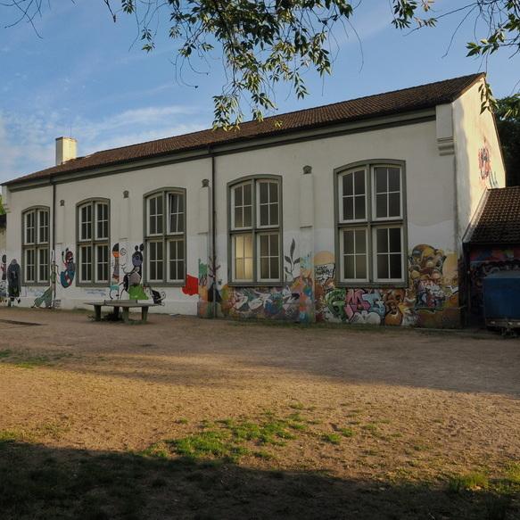 Graffiti-bedeckte Wand eines Geb&auml;udes mit gro&szlig;en Fenstern, umgeben von Gras und Kies. Heller Himmel mit Wolken.