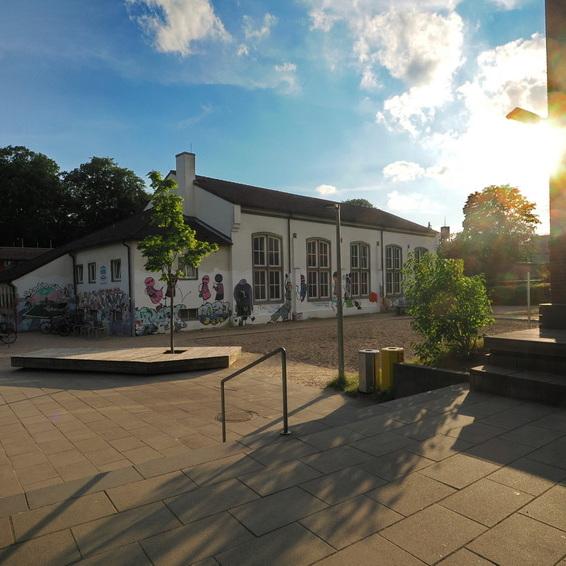 Blick auf ein Schulgeb&auml;ude mit bunten Graffiti und der Sonne, die hinter B&auml;umen und einem modernen Geb&auml;ude scheint.