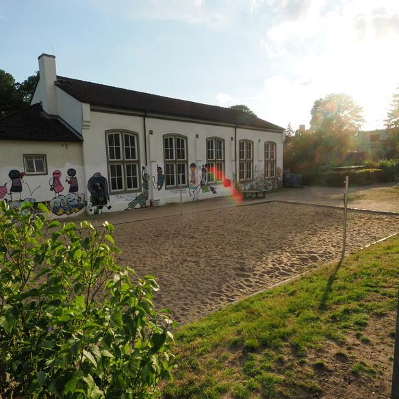 Sandplatz vor einem Geb&auml;ude mit Graffiti an der Wand, umgeben von B&auml;umen und Gras, im Sonnenlicht.