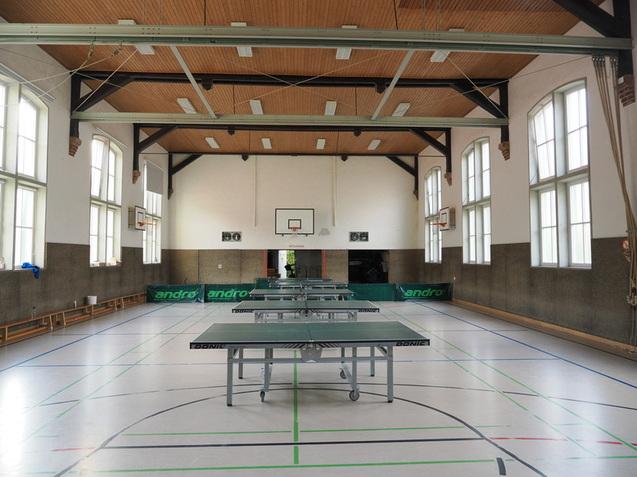 Innenansicht einer Sporthalle mit mehreren Tischtennisplatten und gro&szlig;en Fenstern, Basketballkorb an der Wand.