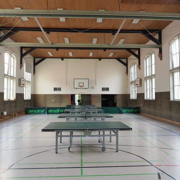 Innenansicht einer Sporthalle mit mehreren Tischtennisplatten und gro&szlig;en Fenstern, Basketballkorb an der Wand.
