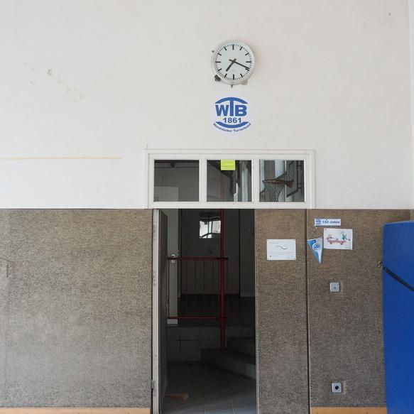 Eingang zu einem Sportbereich mit Uhr und Vereinslogo, Blick auf Treppe hinter einer offenen T&uuml;r.