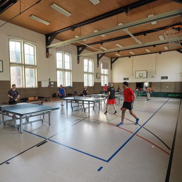 Tischtennistraining in einer Sporthalle mit mehreren Spielern an den Tischen und gro&szlig;en Fenstern im Hintergrund.