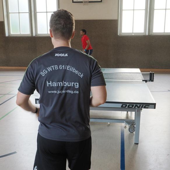 Person in Sporttrikot mit "SG WTB 61/Eilbeck, Hamburg" steht vor einem Tischtennistisch in einer Sporthalle.