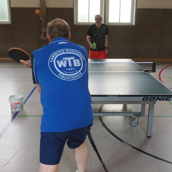Zwei Tischtennisspieler in einem Sportraum, einer schl&auml;gt den Ball, der andere steht am Tisch mit Schl&auml;ger bereit.