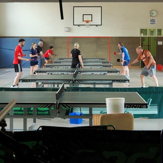 Mehrere Tischtennisspieler trainieren an verschiedenen Tischen in einer Sporthalle mit Basketballkorb im Hintergrund.