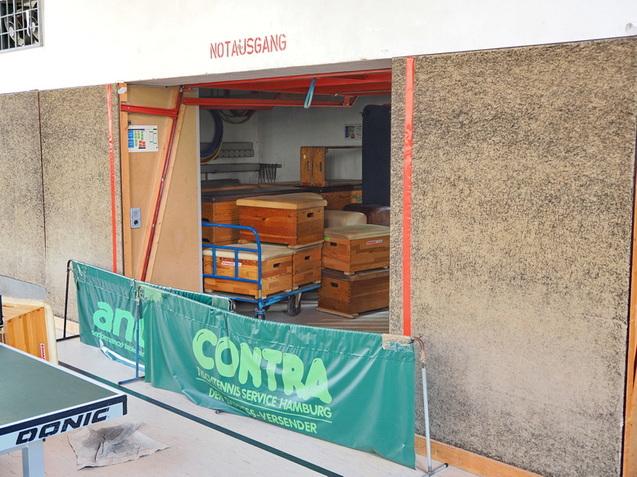 Blick auf einen Notausgang, dahinter Holzregale und -kisten, mit einer gr&uuml;nen Werbebande im Vordergrund.