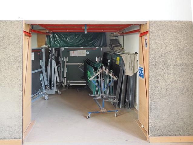 Hintereingang mit gestapelten Tischtennisplatten und St&uuml;hlen in einem Lagerraum.