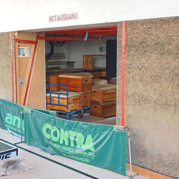 Blick auf einen Notausgang, dahinter Holzregale und -kisten, mit einer gr&uuml;nen Werbebande im Vordergrund.