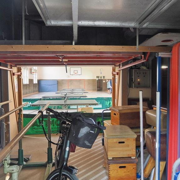 Blick durch eine T&uuml;r auf eine Turnhalle mit Tischtennisplatten, ausgestattet mit verschiedenen Sportger&auml;ten und einer Fahrrad.