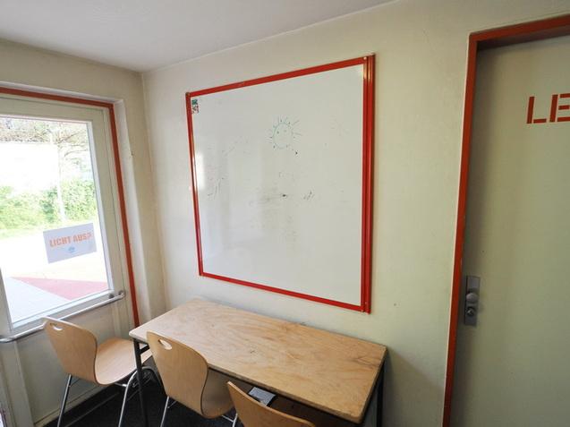 Leere Klasse mit einem Tisch, zwei St&uuml;hlen, einem wei&szlig;en Brett an der Wand und einer T&uuml;r mit der Aufschrift "LEHRER".