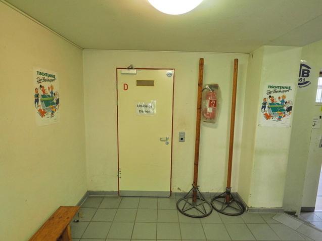 Eingangsbereich mit einer T&uuml;r, zwei Holzst&auml;ndern, einem Feuerl&ouml;scher und Plakaten zum Thema Tischtennis.