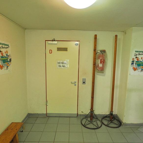 Eingangsbereich mit einer T&uuml;r, zwei Holzst&auml;ndern, einem Feuerl&ouml;scher und Plakaten zum Thema Tischtennis.