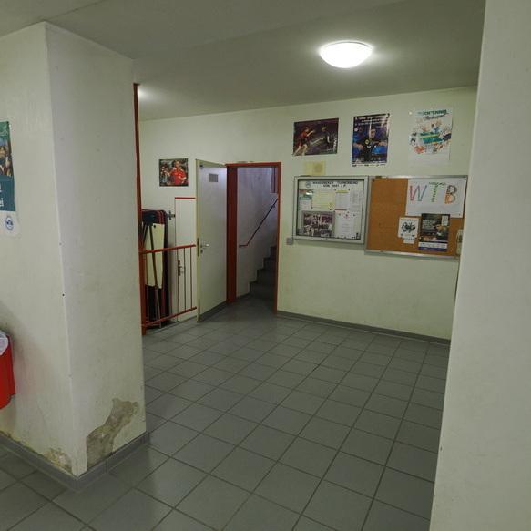 Eingangsbereich mit Treppe, Wandposter und einem Infobrett an einer hellen Wand. M&uuml;llbeh&auml;lter in der Ecke.