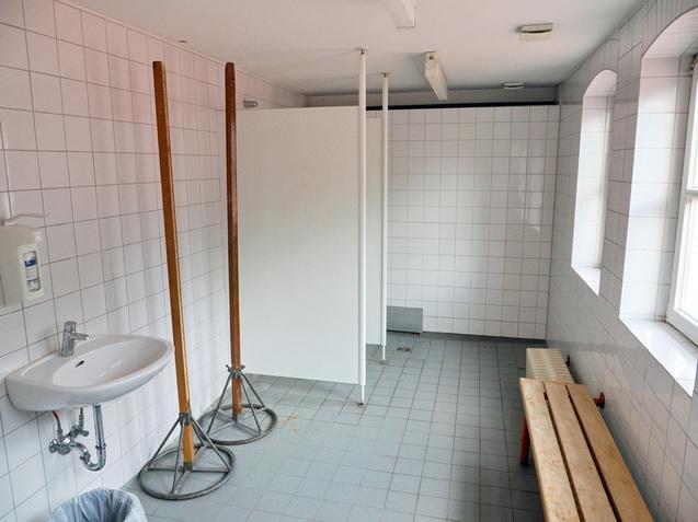 Leere Umkleideraum mit Waschbecken, Holzbank und Trennw&auml;nden auf gefliester Fl&auml;che, beleuchtet von Tageslicht.