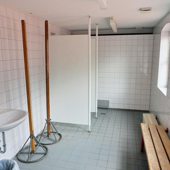 Leere Umkleideraum mit Waschbecken, Holzbank und Trennw&auml;nden auf gefliester Fl&auml;che, beleuchtet von Tageslicht.