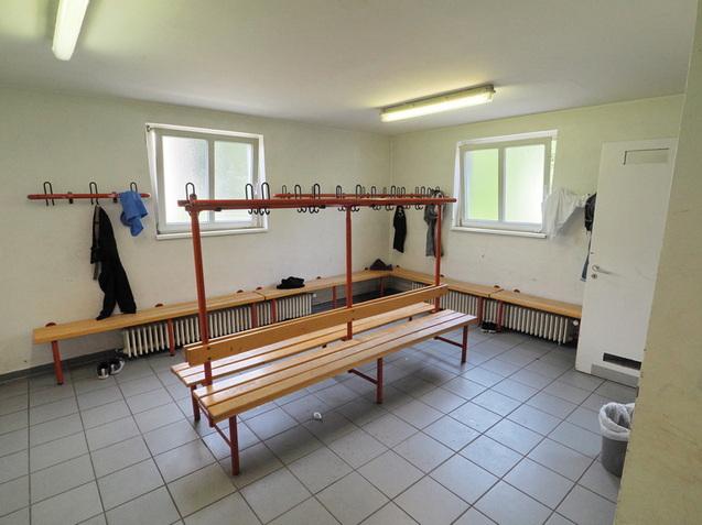 Umkleideraum mit Holzbank, Kleiderhaken an der Wand und Fenster; an den W&auml;nden h&auml;ngen verschiedene Kleidungsst&uuml;cke.