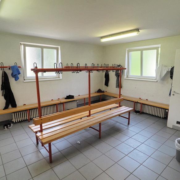 Umkleideraum mit Holzbank, Kleiderhaken an der Wand und Fenster; an den W&auml;nden h&auml;ngen verschiedene Kleidungsst&uuml;cke.
