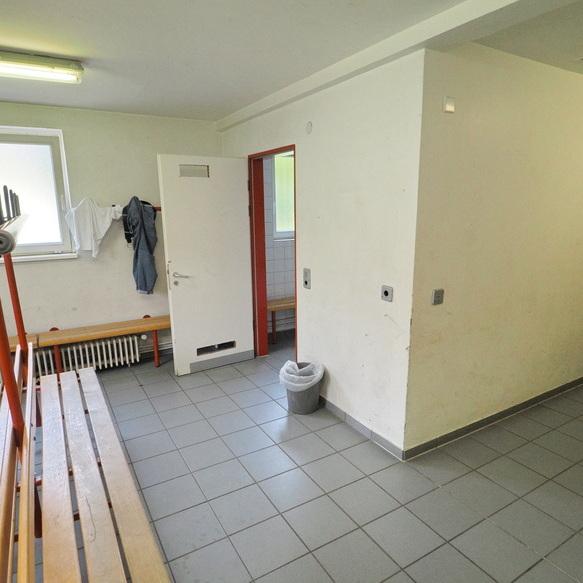 Innenansicht eines Umkleidebereichs mit Sitzelementen, Abstellhaken und Zugang zu einem Badezimmer.