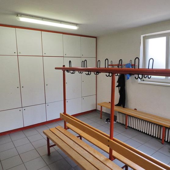 Lockerraum mit wei&szlig;en Schr&auml;nken, zwei Holzbankreihen und Kleiderhaken an einer roten Stange. Fensterlicht f&auml;llt ein.