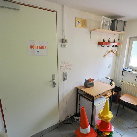 Raum mit einer T&uuml;r, einem Tisch, einem Stuhl, einem Regal und bunten Pylonen, daneben ein Lichtschalter und ein Fenster.