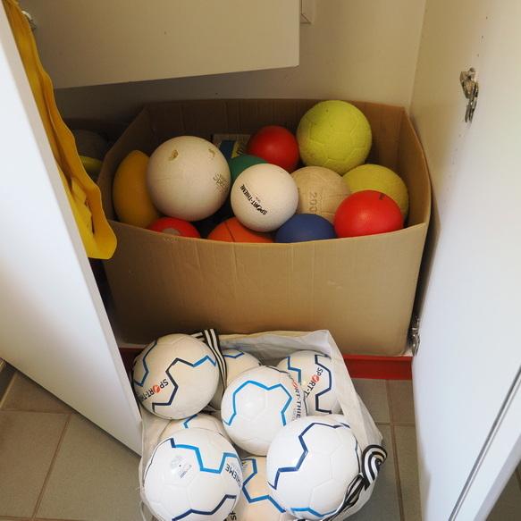 Karton mit verschiedenfarbigen B&auml;llen und eine T&uuml;te mit wei&szlig;en Fu&szlig;ballen in einem offenen Schrank.