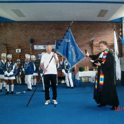 Zwei Männer auf einer Feier: einer hält eine Flagge, der andere spricht mit Mikrofon. Im Hintergrund stehen Personen in Uniform.