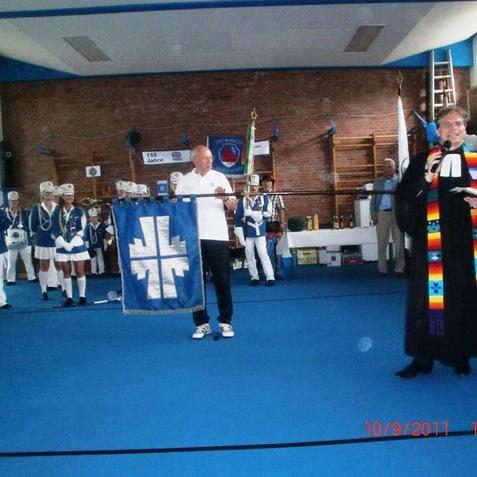 Zwei Männer stehen mit Fahne und Mikrofon in einer Sporthalle, während im Hintergrund Gruppen in Uniform zuschauen.