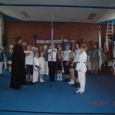 Gruppierung von Kindern in Sportkleidung bei einer Zeremonie, während eine Person eine Fahne hebt.