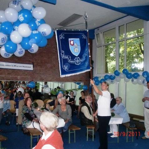 Frau hält Fahne in einem festlich dekorierten Raum mit blau-weißen Ballons und Gästen an Tischen.