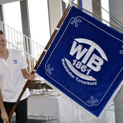 Junger Mann hält eine blaue Fahne mit dem Schriftzug „WTB 1861“ in einem modernen Raum vor einer Treppe.