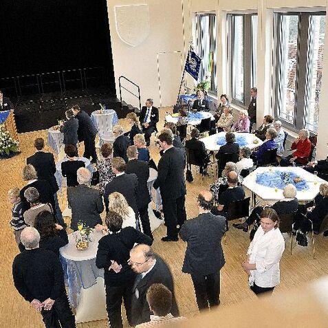 Ein festlicher Saal mit Gästen, die einem Redner an einem Podium zuhören, umgeben von Tischen und Dekoration.