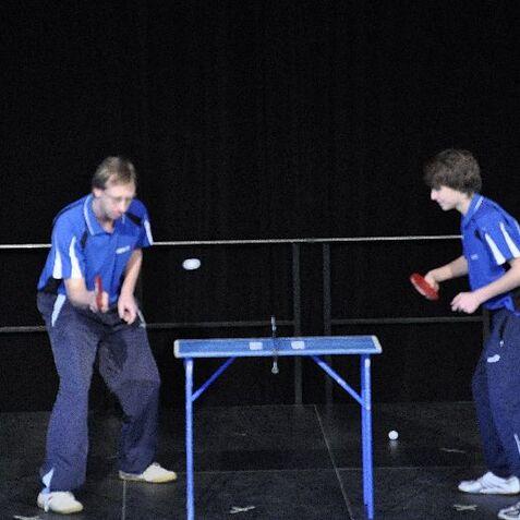 Drei Tischtennisspieler in blauen Trikots spielen an einem Tischtennistisch, während ein Ball in der Luft schwebt.