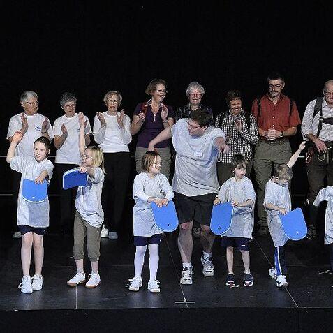 Gruppe von Kindern und Erwachsenen auf einer Bühne, alle mit blauen Schlägern und T-Shirts, applaudierend und lächelnd.