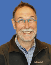 Mann mit Brille und grauem Bart lächelt freundlich vor blauem Hintergrund.