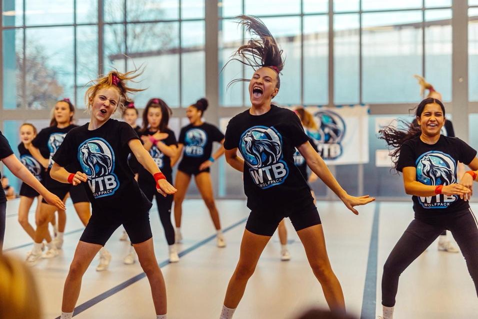 Tanzende Gruppe von Cheerleadern in schwarzen T-Shirts in einer Sporthalle mit großen Fenstern im Hintergrund.