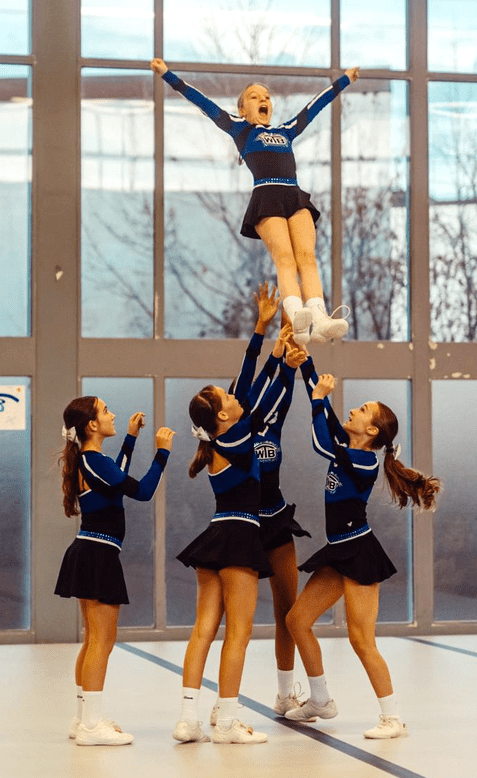 Fünf Cheerleader heben ein Mädchen in die Luft, während sie auf einer senkrechten Fläche in einem Raum trainieren.